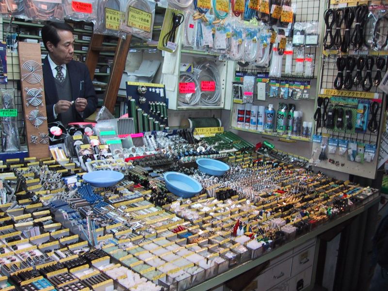 An akihabara shop showing a man dressed in a suit jacket surrounded by small trays of electronic components with cables hanging on the walls.